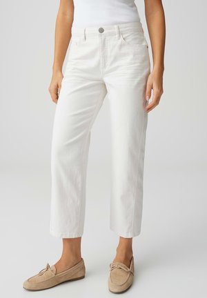 Femme portant un pantalon blanc taille haute à jambes droites avec boutons sur le devant et poches, associé à des mocassins en daim beige.