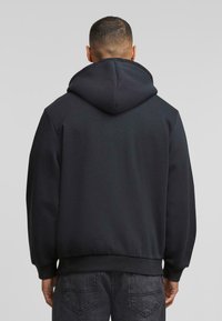 Zwarte hoodie met een relaxte pasvorm, zachte textuur en geribde manchetten. De achterkant heeft een glad design en een grote capuchon.