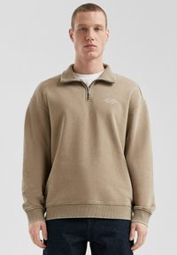 Beige sweatshirt met ritskraag in een ontspannen pasvorm, geribde manchetten en een subtiele tekstdetail op de borst. Zacht materiaal met een gladde textuur.