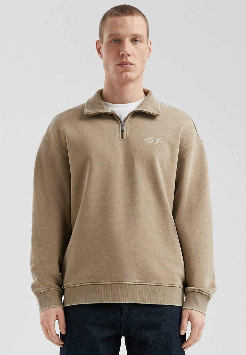 Felpa beige con zip e collo alto, dal taglio rilassato, con polsini a coste e un sottile dettaglio testuale sul petto. Tessuto morbido con una superficie liscia.