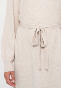 Ljust beige stickad klänning med en mjuk textur, med knytmått vid midjan och ribbade ärmslut. Designen inkluderar långa ärmar och en avslappnad passform.