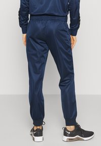Personne portant un pantalon de survêtement bleu marine et des baskets noires Nike, debout dos au spectateur sur un fond uni.