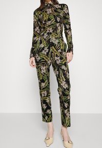 Une combinaison noire avec un motif floral vert et blanc. Manches longues avec des poignets ajustés, jambes amples et texture globalement douce. Chaussures beige clair.