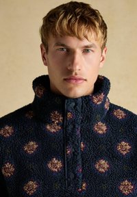 Fleece-Pullover mit hohem Kragen, dunkelblau mit floralem Muster in verschiedenen Lila- und Beigetönen, ausgestattet mit einem Reißverschluss und strukturiertem Stoff.