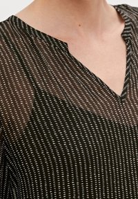 Blouse noire transparente avec un col en V, ornée d'un motif à pois blancs et d'un tissu texturé. La coupe est décontractée avec un tombé fluide.