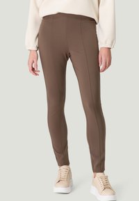 Leggings de sport bruns avec une texture lisse, des détails de couture et une taille haute, associés à des baskets beiges sur un fond neutre.