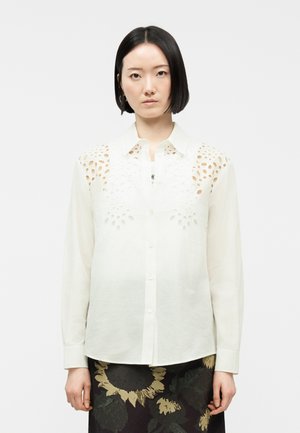 Femeie cu păr scurt negru, purtând o bluză albă cu mâneci lungi și modele decupate pe umeri și o fustă neagră cu imprimeu floral.