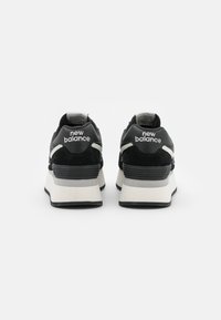 Black suede and leather sneakers with a white midsole, featuring a grey accent and the “New Balance” logo on the heel.