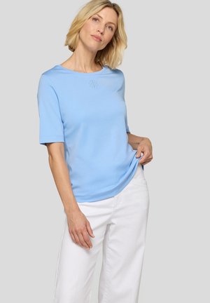 Frau mit kurzen blonden Haaren, die ein hellblaues Kurzarmhemd und weiße Hosen vor einem schlichten hellgrauen Hintergrund trägt.