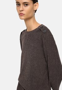 Brązowy prążkowany sweter z okrągłym dekoltem, ozdobiony dużymi czarnymi guzikami na ramieniu i teksturowanym wykończeniem tkaniny.