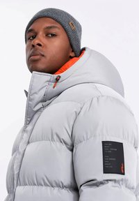 Doudoune isolée gris clair avec intérieur orange, présentant un patch noir sur la manche avec du texte et une capuche avec cordon de serrage.