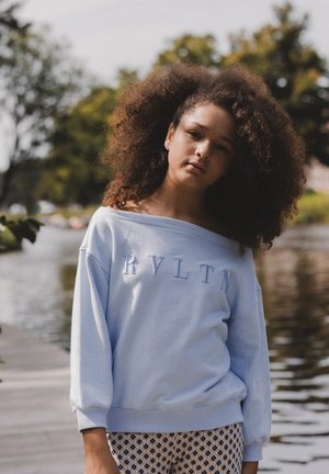 Lichtblauwe sweatshirt met off-shoulder ontwerp en reliëfletters "RVLTN"; gecombineerd met een broek met een patroon van diamanten.