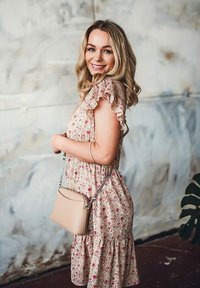 Květinové šaty s volány na rukávech, světle růžová látka a nabraný pas. Béžová kabelka přes rameno se stříbrným řetízkovým popruhem. Pozadí má jemný, neutrální tón.
