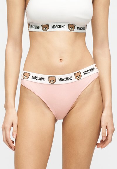 Γυναίκα που φοράει λευκό crop top και ροζ εσώρουχο με λευκές ελαστικές ταινίες που φέρουν το κείμενο Moschino και προσωπάκια αρκουδάκια.