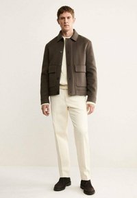 Giacca di lana marrone con grandi tasche frontali, chiusura con bottoni e collo, abbinata a un maglione crema e pantaloni chiari. Scarpa scura.