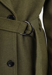 Olivgrön ullrock med tygskärp och silverfärgad rund spänne, svarta knappar och texturerad yta.