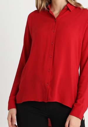 Vrouw met een rood hemd met lange mouwen en knopen, gecombineerd met een zwarte broek, staand tegen een effen lichte achtergrond.