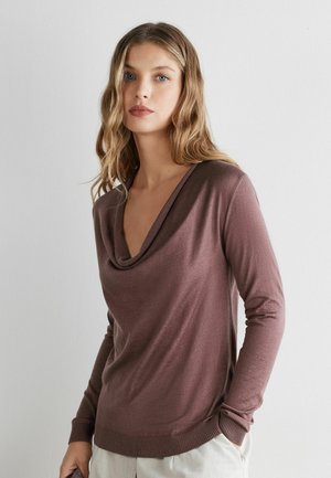 Ung kvinde med bølget blondt hår, iført en lilla langærmet top med krave, stående foran en ensfarvet hvid baggrund.