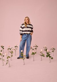 Mujer joven con suéter a rayas y jeans de pie entre rosas rosas en jarrones de vidrio sobre un fondo rosa sólido.