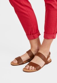 Sandales en cuir marron avec deux sangles horizontales et une sangle réglable à la cheville. Semelle plate et orteils découverts. Portées avec un pantalon rouge retroussé.