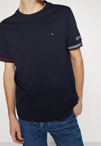 Námořnicky modré bavlněné tričko s krátkými rukávy, na manžetách ozdobené červenými a pruhovanými akcenty a malým logem na hrudi. Viditelné džíny z denimu.