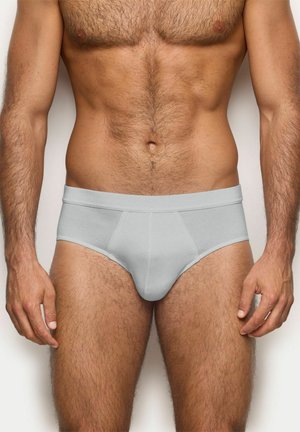 Torso masculino y parte superior de las piernas con calzoncillos gris claro, de pie contra un fondo claro y liso, mostrando el ajuste y estilo de la ropa interior.