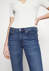 Femme portant un haut en maille blanc à manches courtes et un jean en denim bleu taille haute, avec une main reposant sur la hanche, sur un fond uni.