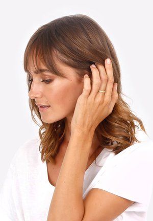 Gold band ring on a hand, worn by a person with medium-length brown hair, textured surface, simple design, against a light background.