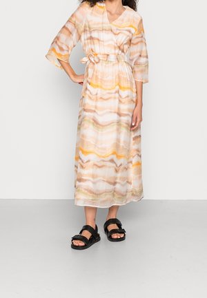 Kvinde iført en lang, flagrende kjole med mønster af bløde orange og beige bølger, bundet i taljen, kombineret med sorte sandaler med åben tå.