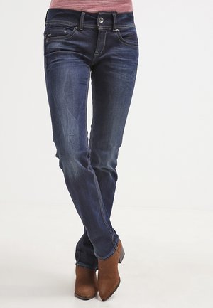 Femme portant un jean bleu foncé ajusté et des bottines marron, debout les jambes croisées devant un fond blanc.