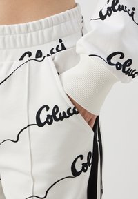 Fehér melegítőnadrág fekete "Colucci" mintával, gumírozott derékkal és bordás mandzsettával. A zseb látható, benne egy kéz. Puha szövet textúra.
