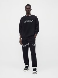Άνδρας που φοράει μαύρο φούτερ Off-White και ταιριαστό παντελόνι με λευκό λογότυπο, στέκεται με το ένα χέρι στην τσέπη μπροστά σε απλό φόντο.
