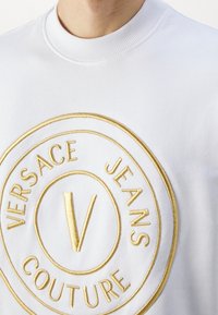 Vit sweatshirt med en stor rund broderad logotyp i guld. Logotypen innehåller texten "VERSACE JEANS COUTURE" och ett stiliserat "V."
