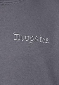 Szary sweatshirt z haftem "Dropsize" w białym, gotyckim stylu. Materiał wydaje się miękki, o gładkiej fakturze i klasycznym kroju z okrągłym dekoltem.
