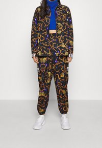 Svart mönstrad två-delad outfit med djärva orange kedjor och metalliska accent, parat med en blå ribbad tröja och vita sneakers.