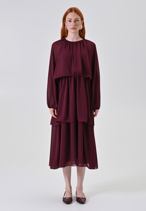 Robe bordeaux à plusieurs niveaux avec des manches longues et un col haut, présentant un tissu fluide et transparent ainsi qu'un design à volants, complété par des chaussures brillantes.