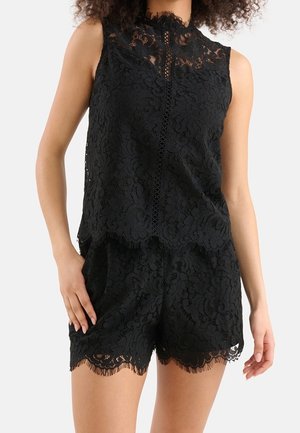Sleeveless black lace top with high neck and matching lace shorts, worn by a standing person with hands relaxed at sides.