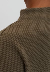 Světle zelený svetr s texturou a geometrickým vzorem, s vysokým límcem a surovými okraji na výstřihu a ramenním švu.