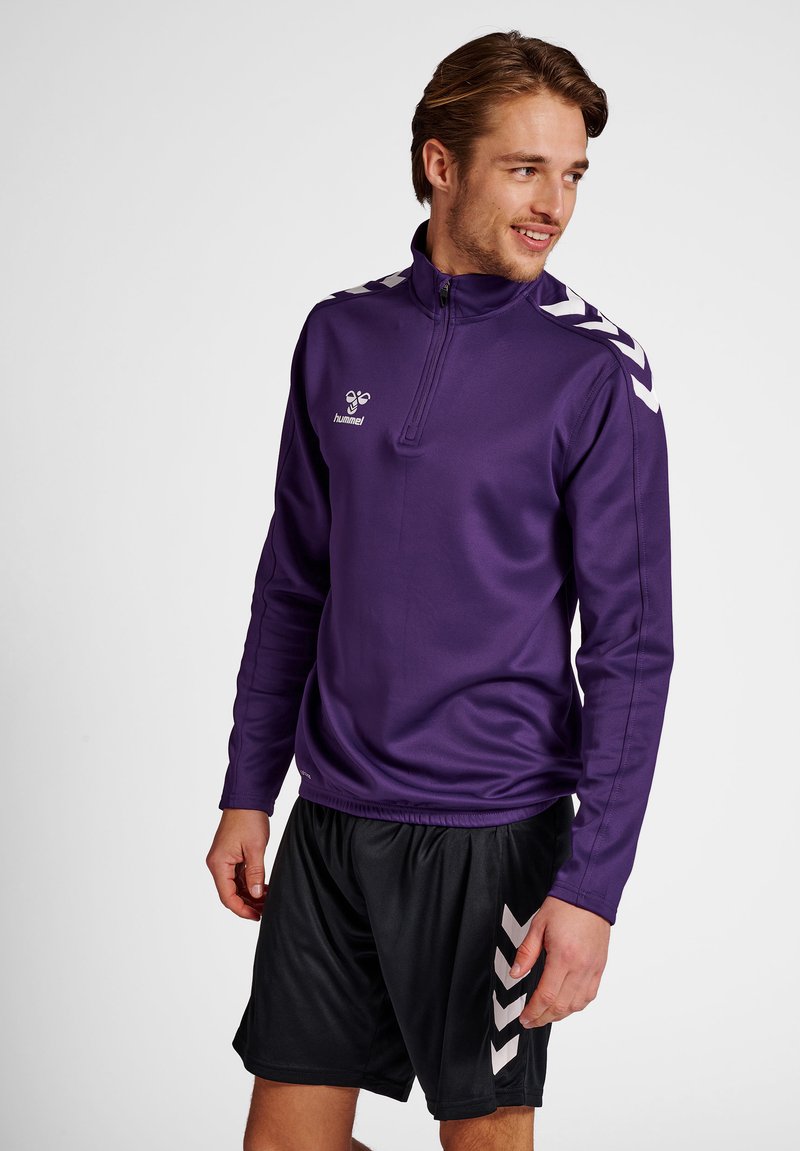 Fioletowy sportowy polar z suwakiem na ćwierć długości, z białymi paskami na ramionach i logo na klatce piersiowej, w połączeniu z czarnymi szortami.