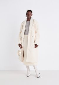 Manteau en laine double boutonnage crème, haut gris ajusté, jupe blanche texturée, bottes à talons blanches brillantes et un petit sac à main crème avec des volants.