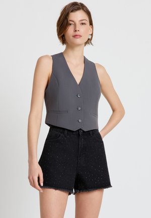 Femme aux cheveux courts portant un gilet gris sans manches boutonné et un short noir taille haute avec de petites clous et des bords effilochés.