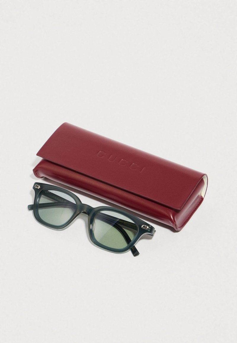 Green-tinted rectangular sunglasses with dark green frames beside a closed maroon leather Gucci case on a light surface.