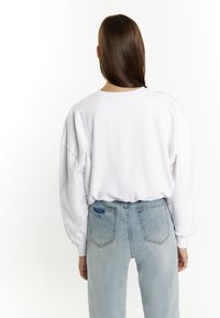 Witte oversized sweatshirt met verlaagde schouders, gecombineerd met lichtblauwe versleten jeans met zichtbare achterzakken en een lichte rafeling.