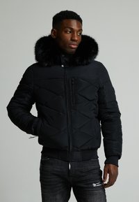 Giacca a vento nera con colletto in pelliccia sintetica, design a rombi imbottito, chiusura con zip e bottoni a pressione, e polsini a coste, abbinata a jeans scuri.
