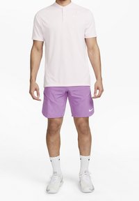 Man i ljusrosa kortärmad polo shirt, lila träningsshorts, vita ankelsockor och vita Nike sportskor, stående mot vit bakgrund.