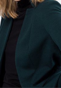 Veste bleu sarcelle foncé en tissu texturé, présentant une coupe ajustée et une ouverture sur le devant, portée sur un col roulé noir à manches longues.