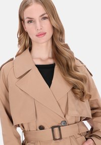 Beige trenchcoat med brede revers, dobbeltradede knapper og et bælte. Stoffet ser glat ud med en struktureret silhuet. Sort top under.