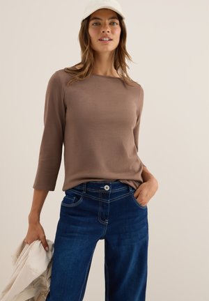 Maglietta marrone a maniche lunghe con scollatura ampia, abbinata a jeans di denim blu scuro con cuciture in evidenza e chiusura a bottone.