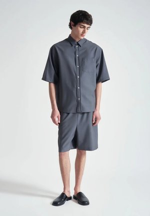 Giovane uomo che indossa una camicia a maniche corte grigia scura e pantaloncini abbinati con scarpe slip-on nere, in piedi davanti a uno sfondo semplice.