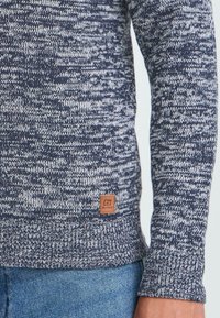 Närbild av ärmen och fållen på en blå och vit stickad tröja med lädermärke, buren över ljusblå denimjeans.